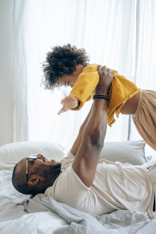 Side view bearded black father in white casual shirt resting on cozy bed with white sheets and lifting up happy little boy in orange pajama after awakening in light bedroom