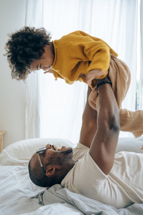 Side view black man wearing casual outfit and eyeglasses relaxing on comfy bed and lifting up cheerful son in cozy pajama against window in light bedroom during sunny morning