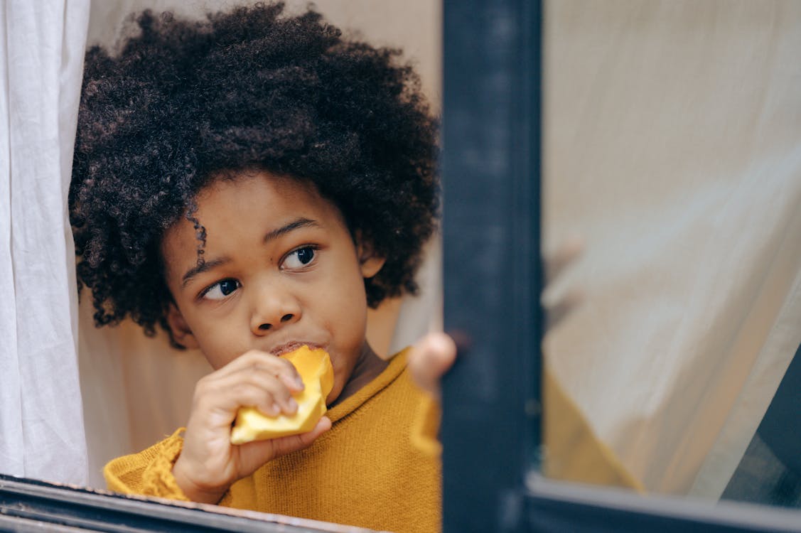 Curious little African American boy with curly hair wearing yellow casual clothes eating sweet exotic fruit and looking out opened window while standing against white curtains