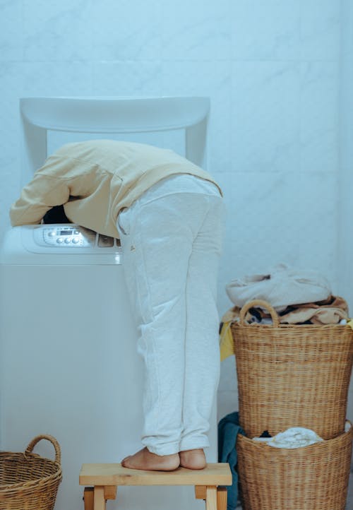 Free Back view full length barefoot faceless ethnic child in casual outfit standing on stool and taking clothes out of washing machine with top loader while keeping head inside of washer Stock Photo