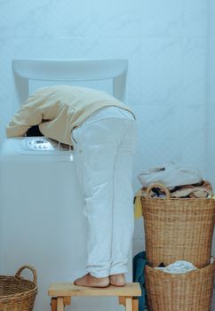 一个穿着休闲装、赤脚不露面的民族儿童站在凳子上，从顶部装载的洗衣机里取出衣服，同时将头保持在洗衣机内