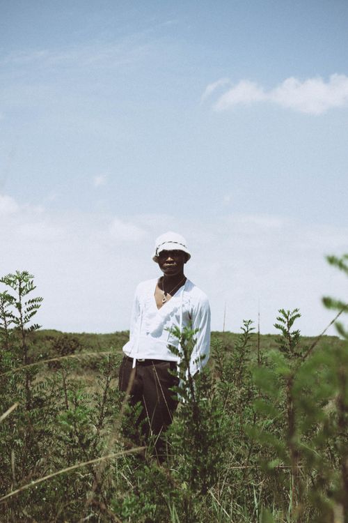 Free A fashionable man in Nairobi's natural setting, wearing a white top and hat, surrounded by greenery. Stock Photo
