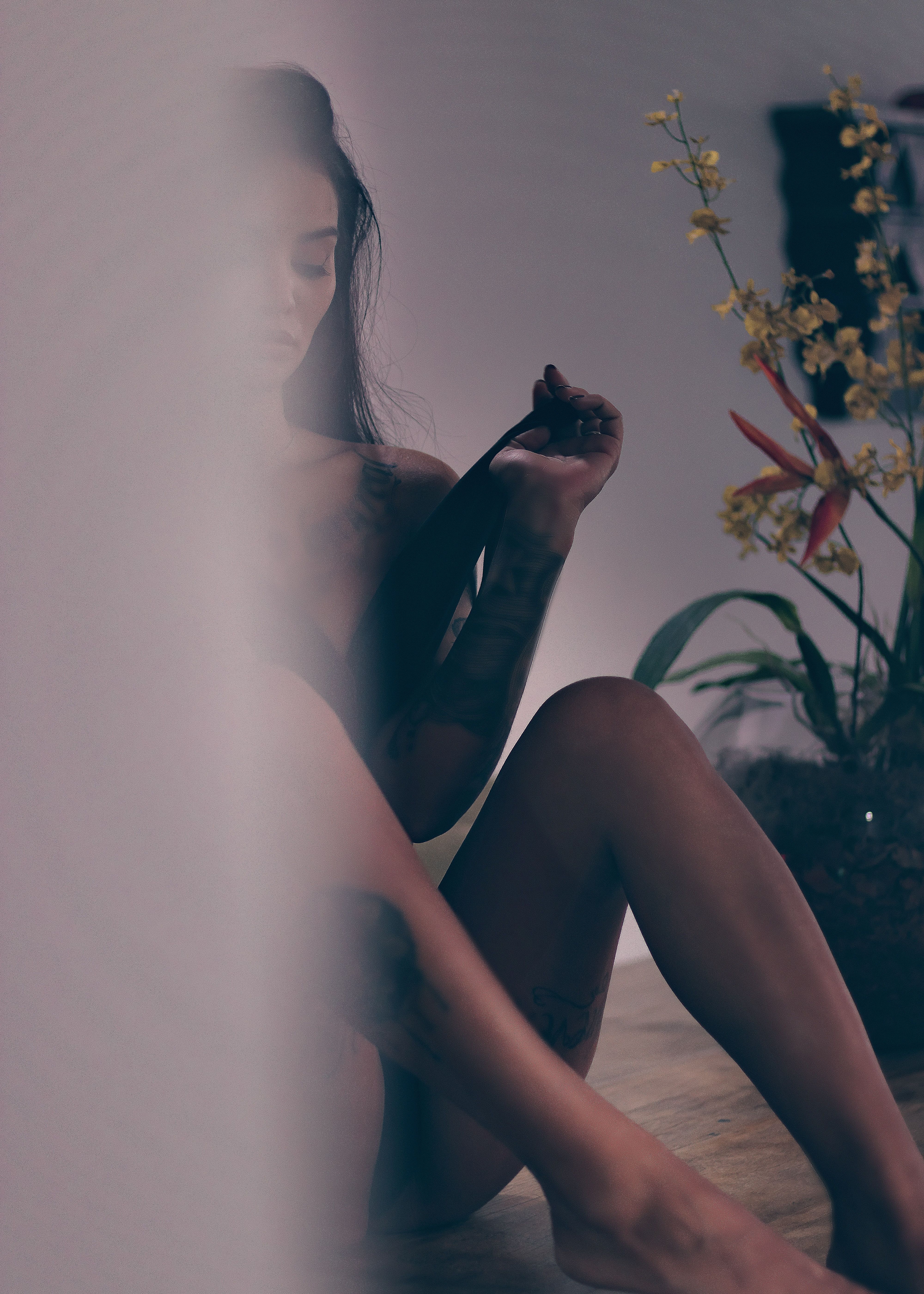 Sultry portrait of a tattooed woman sitting indoors with soft lighting and floral decor.