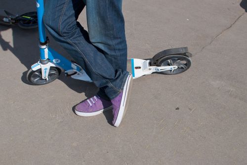Free Close-up of a person in jeans and purple sneakers leaning on a blue kick scooter on a sunny day. Stock Photo