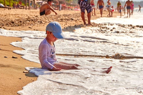 A Kid Wearing Cap and White Long Sleeves Sitting on the Shore of a Beach