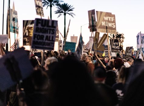 A diverse crowd gathers outdoors holding protest banners and signs at dusk.