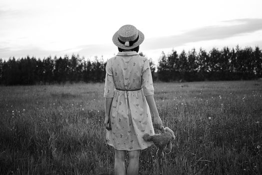 Black and white back view of faceless female in hat with soft toy standing on grass lawn in front of trees under cloudy sky