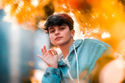 From below of confident young millennial with dark short hair in stylish hoodie chilling in autumn park and touching neck chain