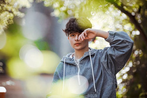 Free Serious young millennial in trendy outfit rubbing eye and looking at camera while standing near green trees in park Stock Photo