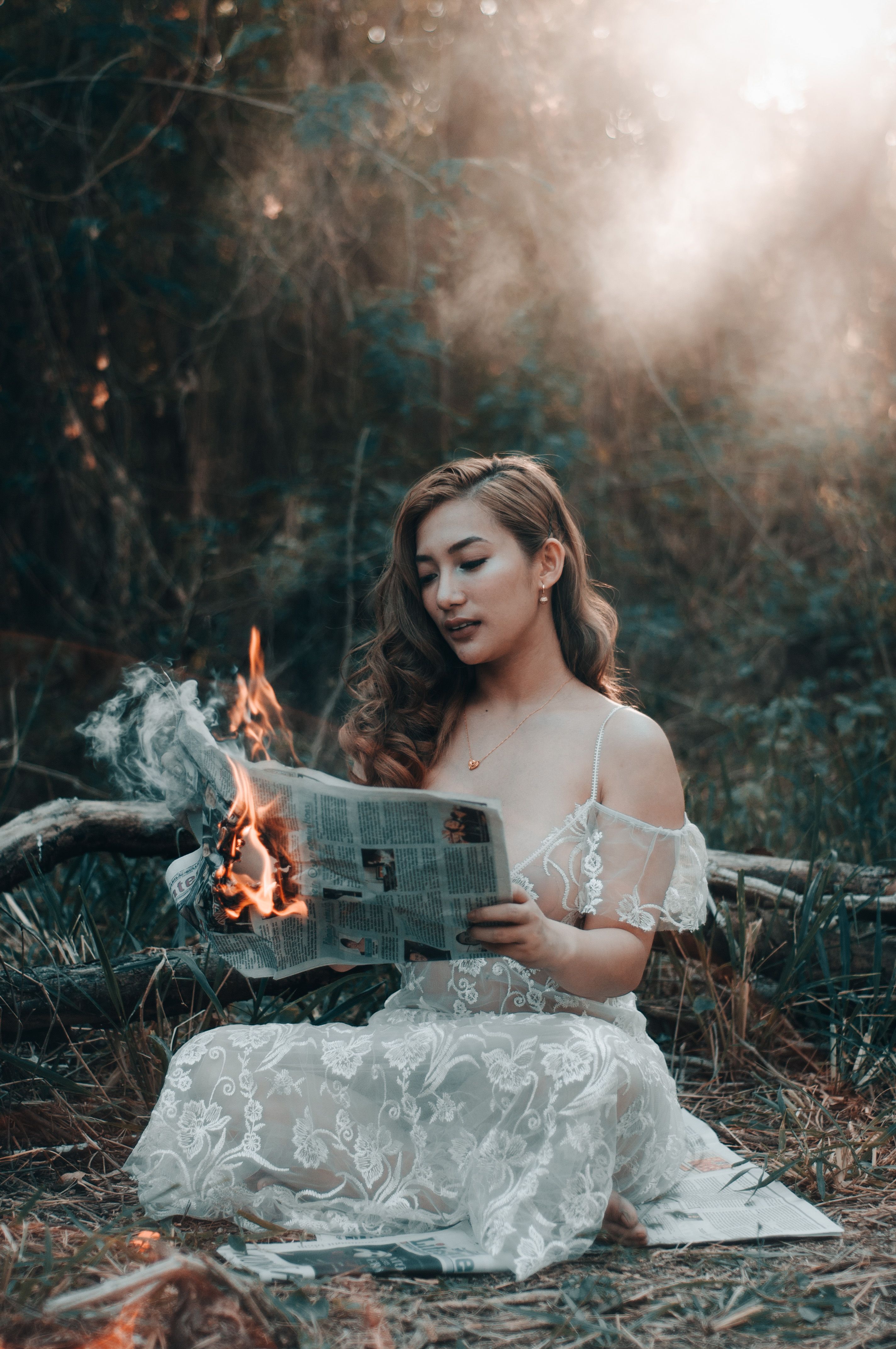 一位身着白色连衣裙的女子在神秘的森林中阅读燃烧的报纸