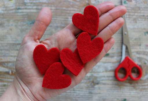 一只手握着红色毛毡心形切口，它位于木制表面上，非常适合制作情人节工艺品