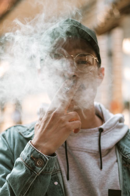 Confident young ethnic male in trendy outfit and eyeglasses hidden behind smoke of cigarette on street