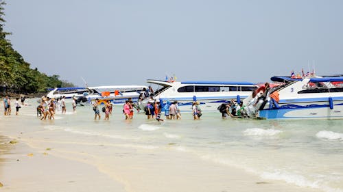 Crowd of anonymous tourists walking on sandy ocean beach near modern cruise boats moored on coast against cloudless blue sky