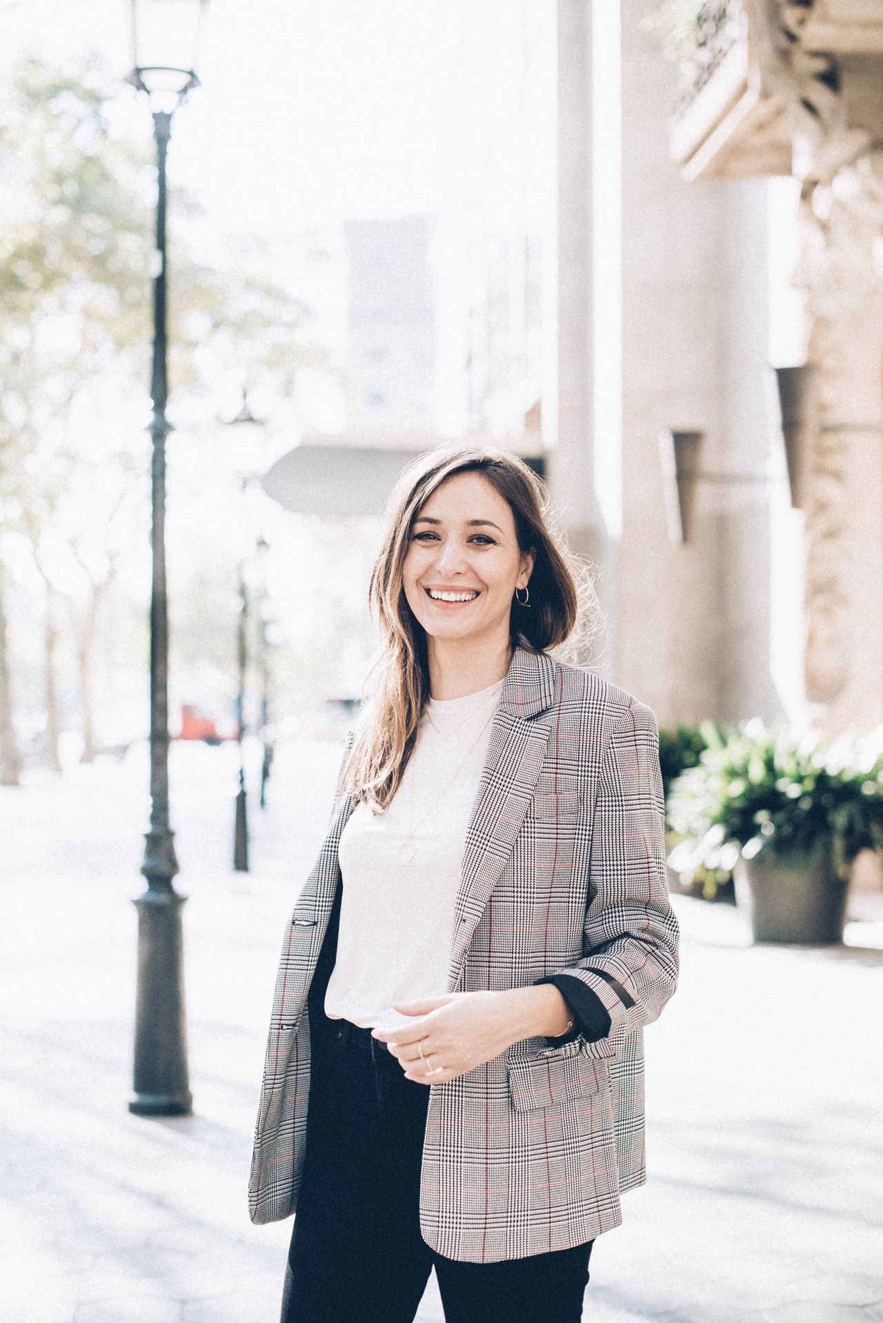 Smiling woman in a gray plaid blazer, white top, and black trousers standing on a sunny city street.