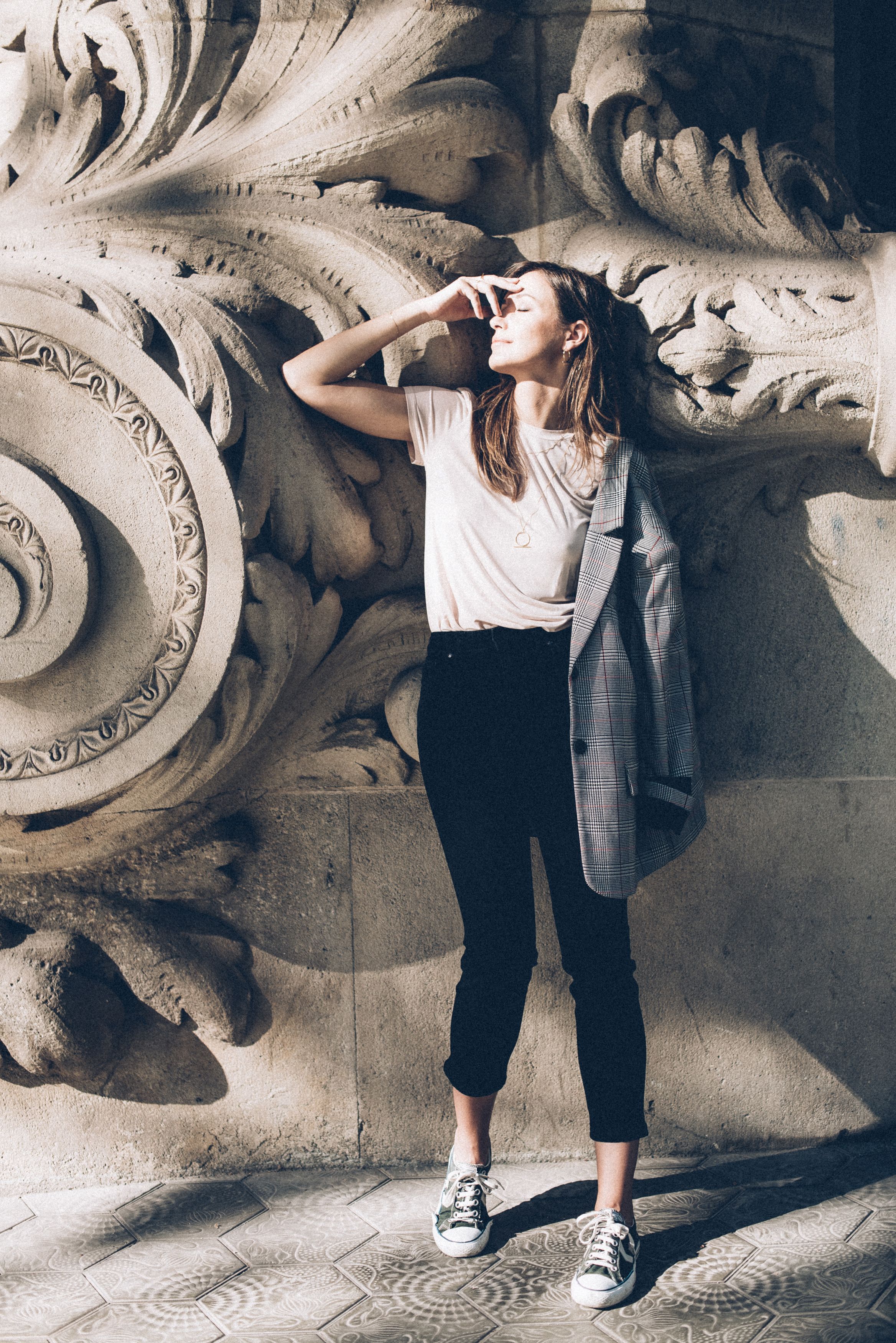 Fashionable young woman in a blazer and white shirt, basking in sunlight against ornate architecture.