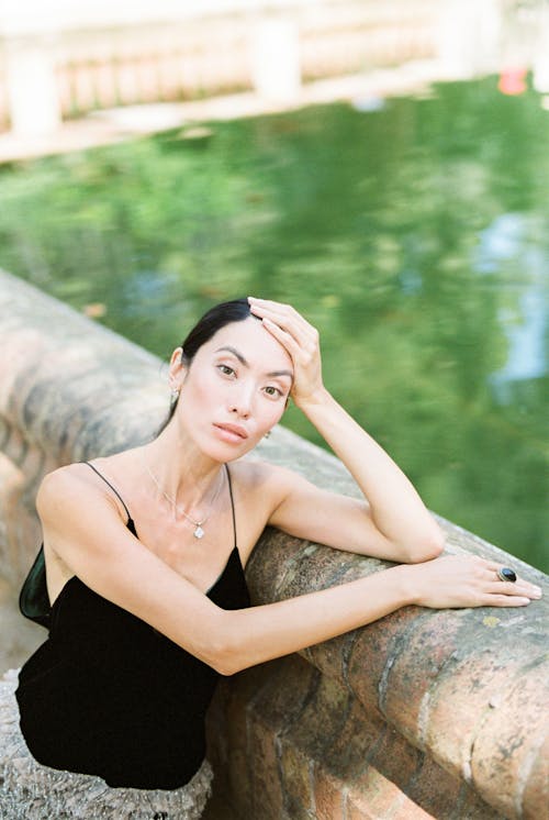 A Woman in Black Spaghetti Strap Top Leaning on Concrete Railing with Her Hand on Her Head