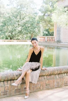 A fashionable woman in a black dress sitting by a pool, showcasing elegance and style in a serene outdoor setting.