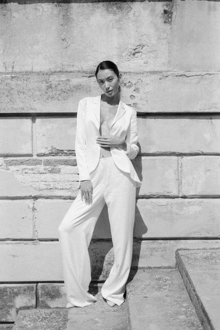 Stylish woman in white suit posing against a textured wall in a monochrome setting.