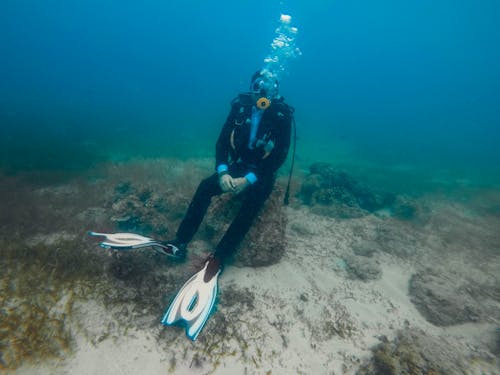 Fit anonymous frogman in diving dress and flippers sitting on stone with hands together under ocean water with sand and seaweed on bottom and bubbles above oxygen mask