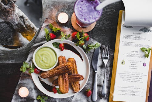 Free From above of delicious Spanish churros with green dip sauce garnished with fresh strawberries and melted chocolate near glass of blueberry milkshake and menu in restaurant Stock Photo