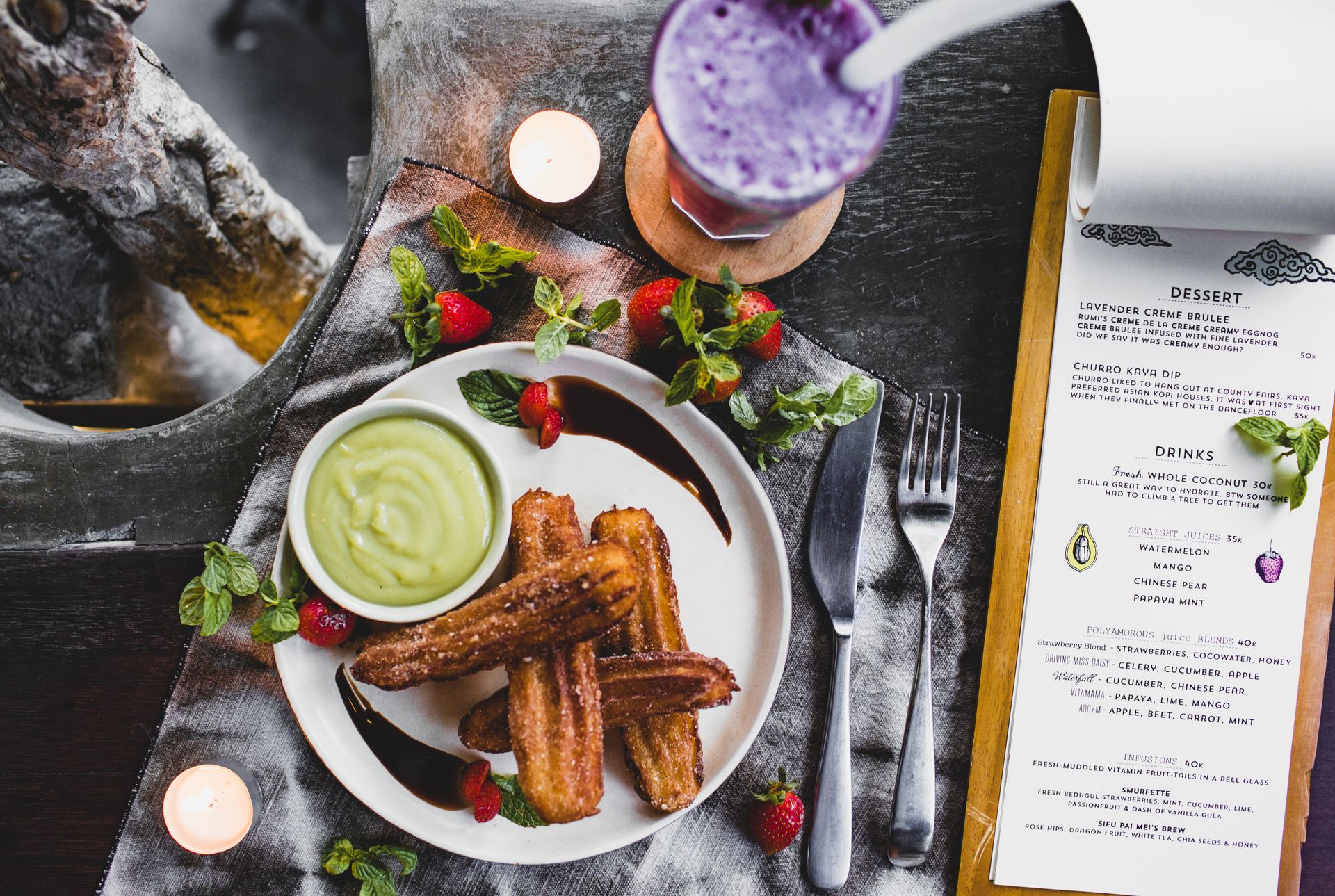 From above of delicious Spanish churros with green dip sauce garnished with fresh strawberries and melted chocolate near glass of blueberry milkshake and menu in restaurant