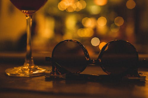 Composition of glass with refreshing drink on thin leg near modern sunglasses for women in plastic frame on wooden table in restaurant at night