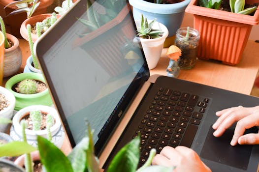 Hands typing on a laptop surrounded by potted plants in a cozy indoor setting.