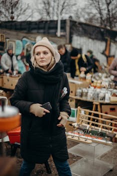A woman in winter attire holding a phone, navigating through a vibrant outdoor flea market.