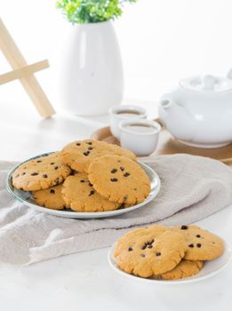 Plate of fresh chocolate chip cookies accompanied by white teapot and potted plant.