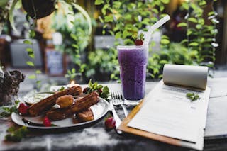 Glass of yummy blueberry milkshake with straw near plate of churros with fresh strawberries and menu with cutlery on table in front of climbing plant in restaurant