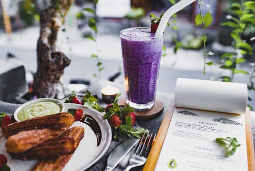 Free From above of glass of lavender creme brulee garnished with strawberry and plate of churro with avocado sauce placed on table with knife and fork and menu and candle against window decorated with plants Stock Photo