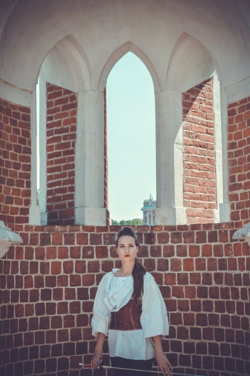 Free Concentrated young lady in old fashioned costume with epee in hand standing against stone wall of ancient castle with arched windows and looking at camera Stock Photo