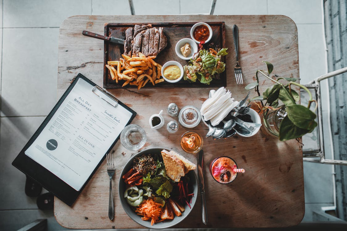 Free Top view of wooden table with salad bowl and fresh drink arranged with tray of appetizing steak and french fries near menu in cozy cafe Stock Photo