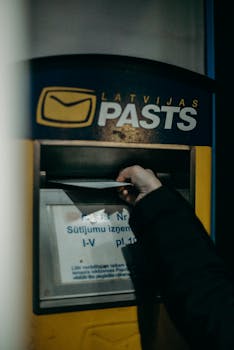 A hand is seen dropping a letter into a Latvian postal box at night.