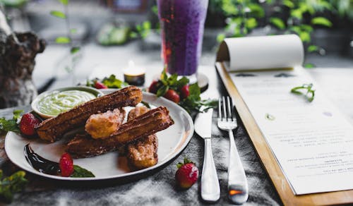 Crispy sweet churros served with sauce and strawberries and placed on table near menu and fresh blueberry smoothie