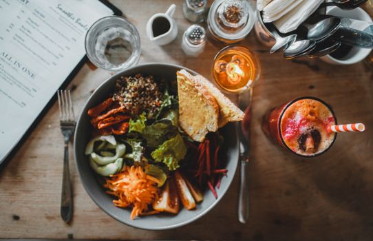Top view of healthy salad with assorted vegetables and toasted bread placed on wooden table with exotic fresh drink and cutlery