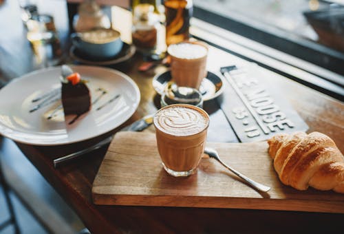 Free From above of glass of cold coffee drink served with fresh crunchy croissant on wooden board near plate with chocolate cake in cozy cafe Stock Photo