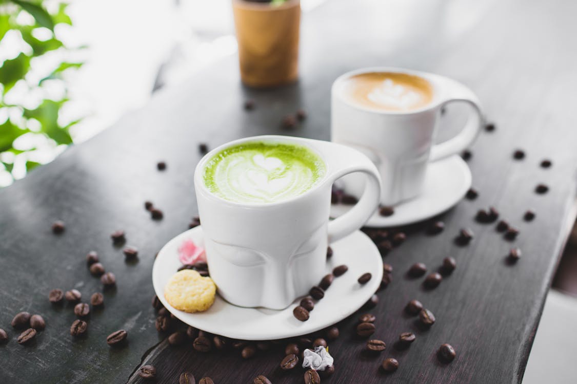 Free From above of refreshing tasty matcha green tea latte and cappuccino in creative face shaped mugs placed on wooden table with scattered coffee beans Stock Photo