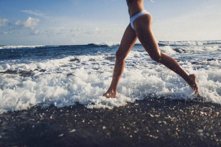 Crop Slim Woman Running On Beach Through Foamy Waves Of Ocean