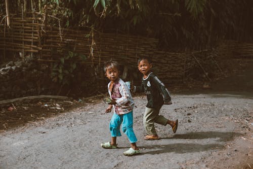 Full body of little ethnic children walking on dirty path near green plants in village on sunny day