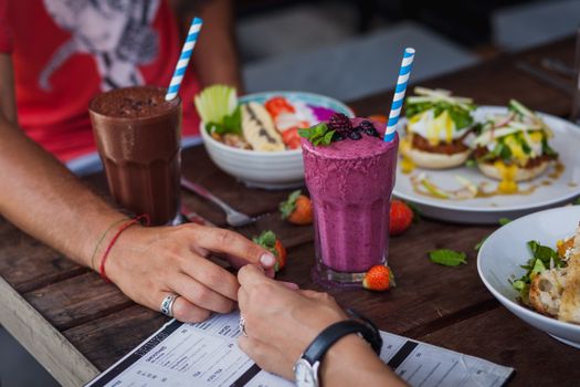 Crop unrecognizable man and female holding hands while having lunch with appetizing dishes and smoothies in tropical cafe