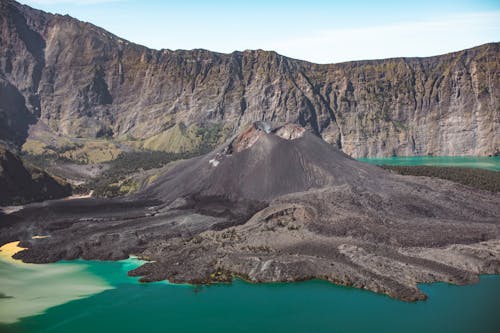 Breathtaking scenery of majestic Rinjani volcano located amidst crater lake with clean green water and surrounded with rocky mountain range against cloudless blue sky