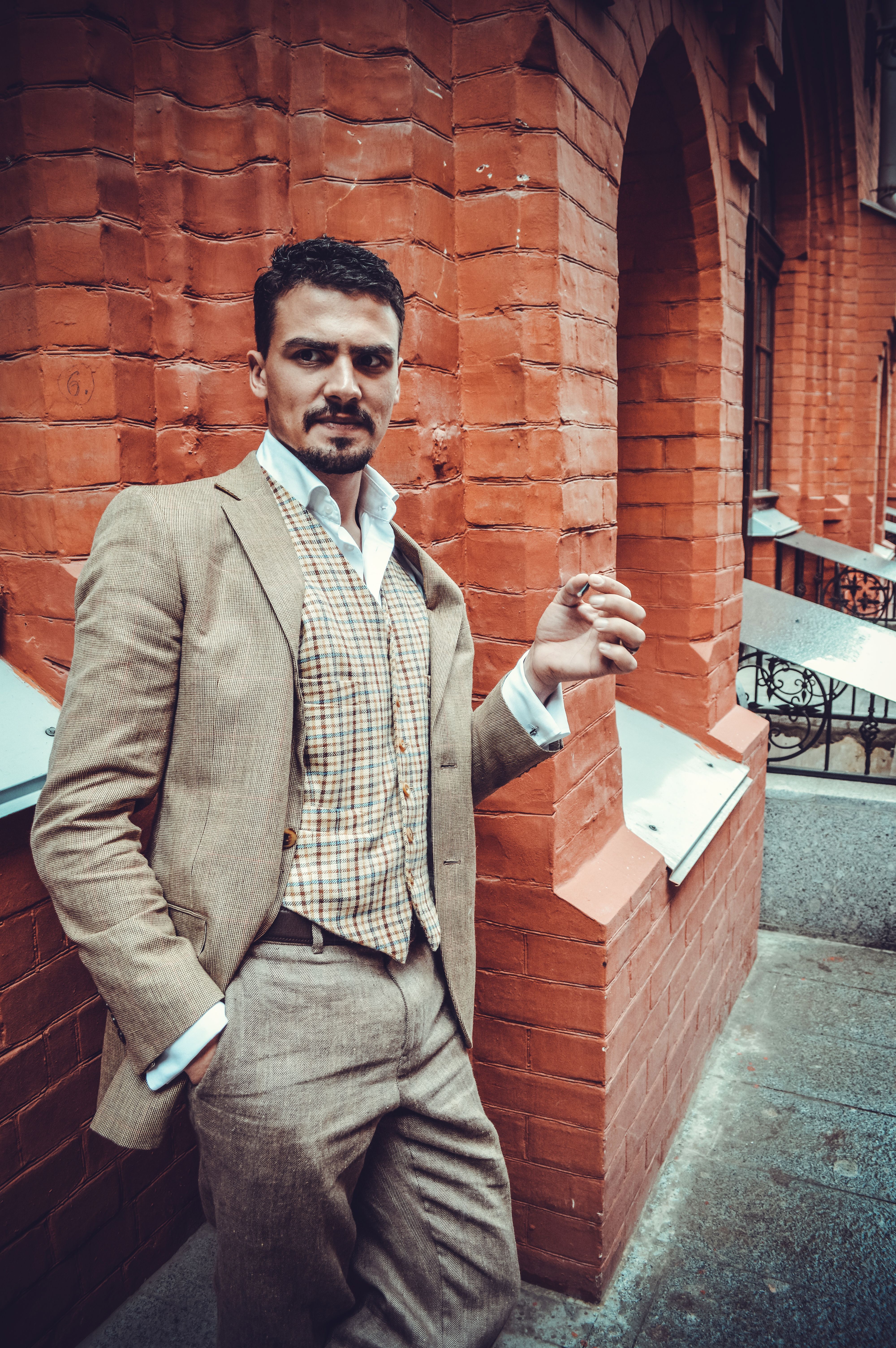 Confident male with black hair and mustache wearing check vest leaning on brick wall while standing with hand in pocket on street