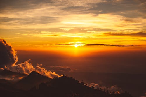 Majestic scenery of bright evening sun illuminating cloudy sky in vivid red and orange colors against silhouette of mountain range covered with trees