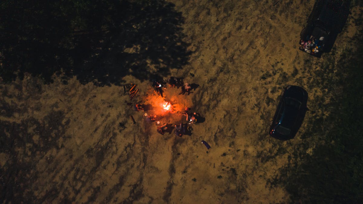 Drone view of group of friends spending time together by flaming bonfire in woodland at night