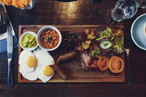Top view of delicious fried eggs and vegetables with sausages and beans on wooden tray with fork and knife
