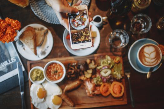 Top view of crop unrecognizable person taking photo of satisfying breakfast and desert with coffee on mobile phone while sitting at table