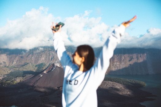一位身穿白色毛衣的年轻女性站在山边，手里拿着手机，面容不清晰
