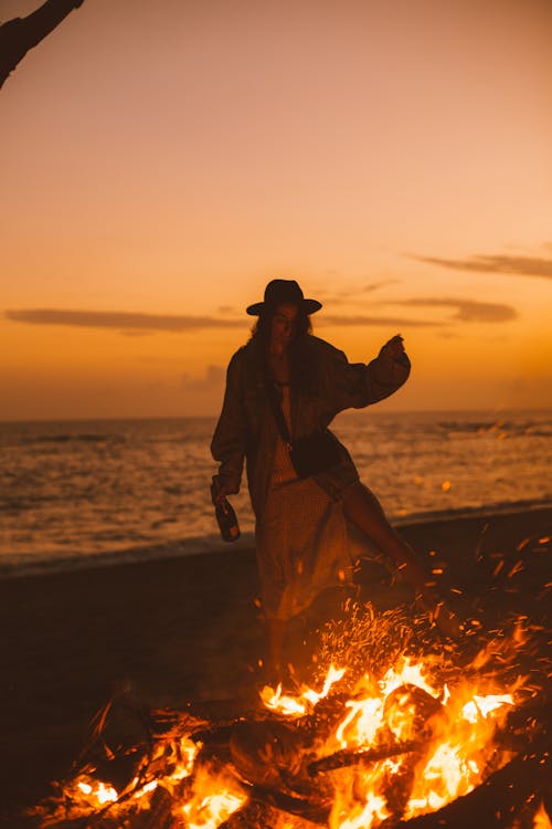 Young female with hat standing near sea and enjoying fire in evening time