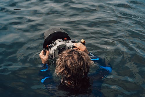 Back view of unrecognizable diver in wet suit taking picture on professional equipment for undersea imaging while swimming in sea